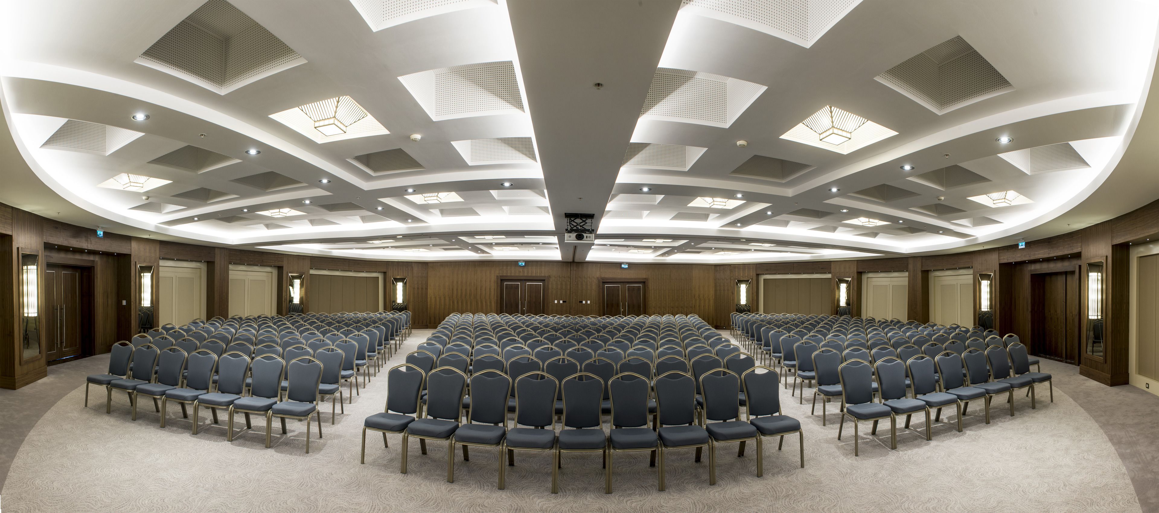 A conference room with rows of chairs, a high ceiling with recessed lighting, and wooden paneling on the walls.
