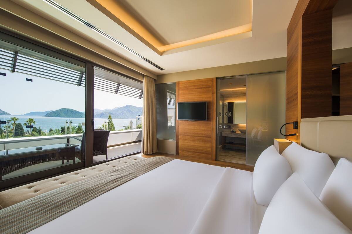 A modern hotel room with a large bed, a balcony with a view of the sea, and a TV mounted on the wall.