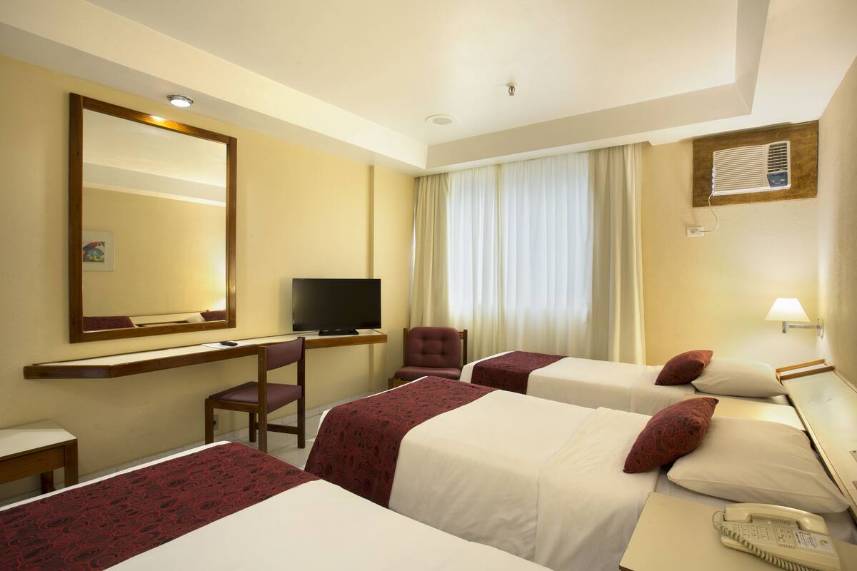 Ein Hotelzimmer mit drei Betten, einem Schreibtisch mit Fernseher, einem Sessel, einem Telefon und einem Vorhangfenster.