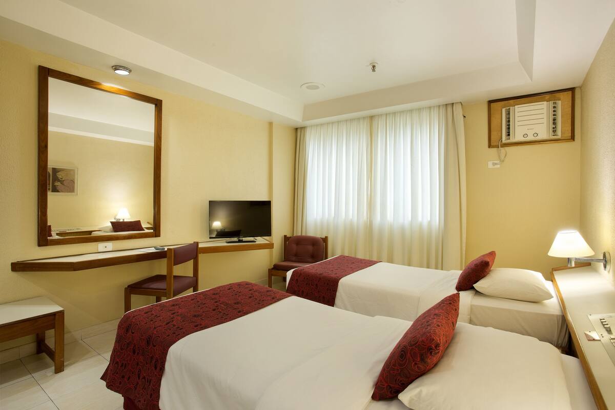 Ein Hotelzimmer mit zwei Betten, einem Schreibtisch, einem Stuhl, einem Fernseher und einem Fenster mit Vorhängen.