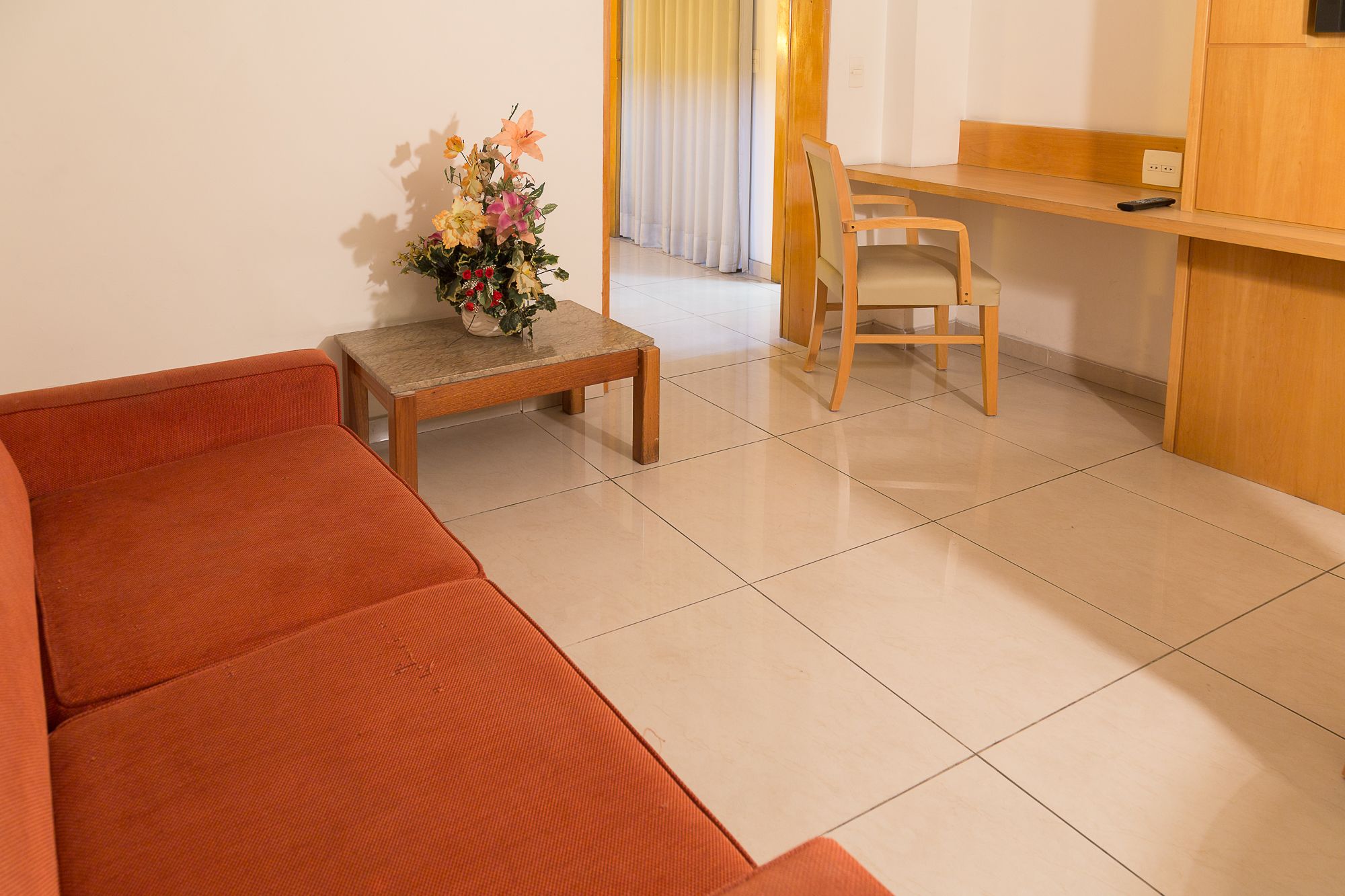 A room with a sofa, a small table with a vase of flowers, a wooden chair, and a desk with a remote control.