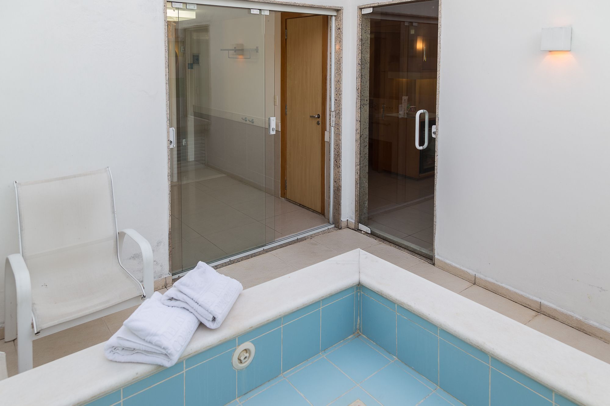 Een hoek van een kamer met een blauw betegeld bad, een witte stoel en een glazen deur die naar een andere kamer leidt.