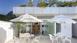 Une terrasse avec des chaises et des tables en plastique blanc, deux parasols blancs et, au fond, du matériel d’exercice.