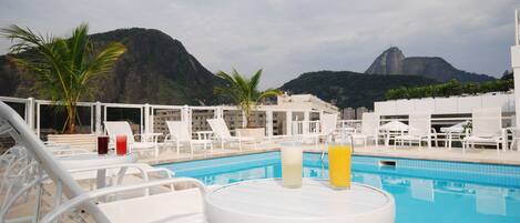 A poolside area with lounge chairs and a table with drinks, overlooking a mountain and a building.
