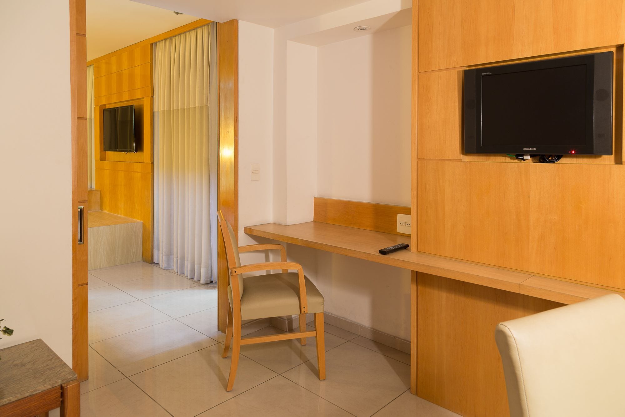 A modern room with a wooden desk, a chair, a television mounted on the wall, and a window with curtains.