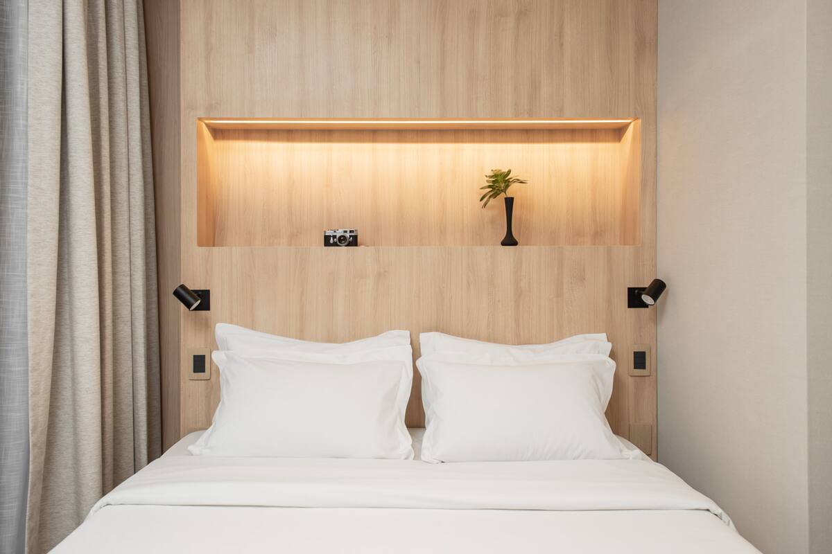 Une chambre d’hôtel avec une tête de lit en bois, une table de chevet sur laquelle se trouve un appareil photo, une petite plante en pot et deux lampes de chevet.