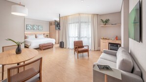 A modern hotel room with a bed, a sofa, a dining table, and a kitchen area.