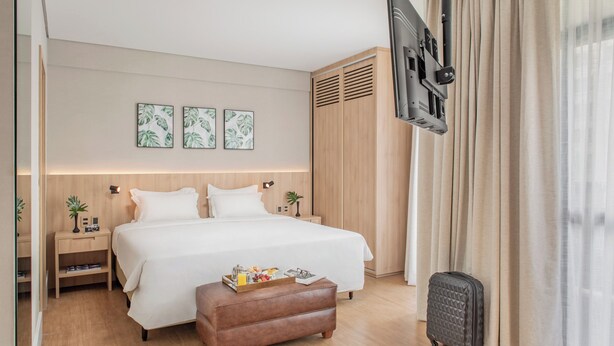 A hotel room with a large bed, a flat-screen TV mounted on the wall, a wooden wardrobe, and a small bedside table with a lamp.