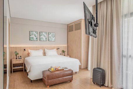 A hotel room with a large bed, a flat-screen TV mounted on the wall, a wooden wardrobe, and a small bedside table with a lamp.