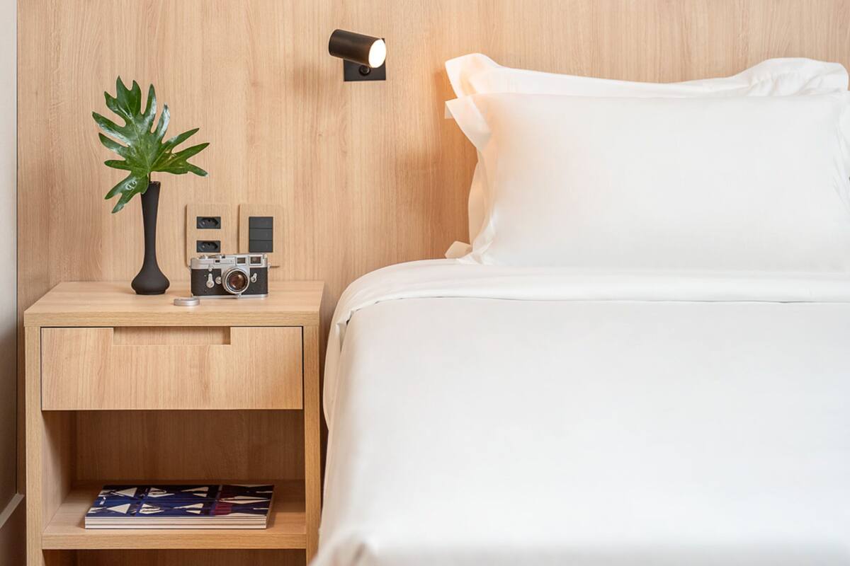 Un lit bien fait, avec du linge de lit blanc, une table de chevet en bois ornée d’une petite plante en pot, et deux reproductions botaniques encadrées accrochées au mur.