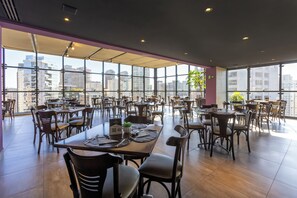 A spacious dining area with multiple tables and chairs, large windows offering a city view, and a modern interior design.
