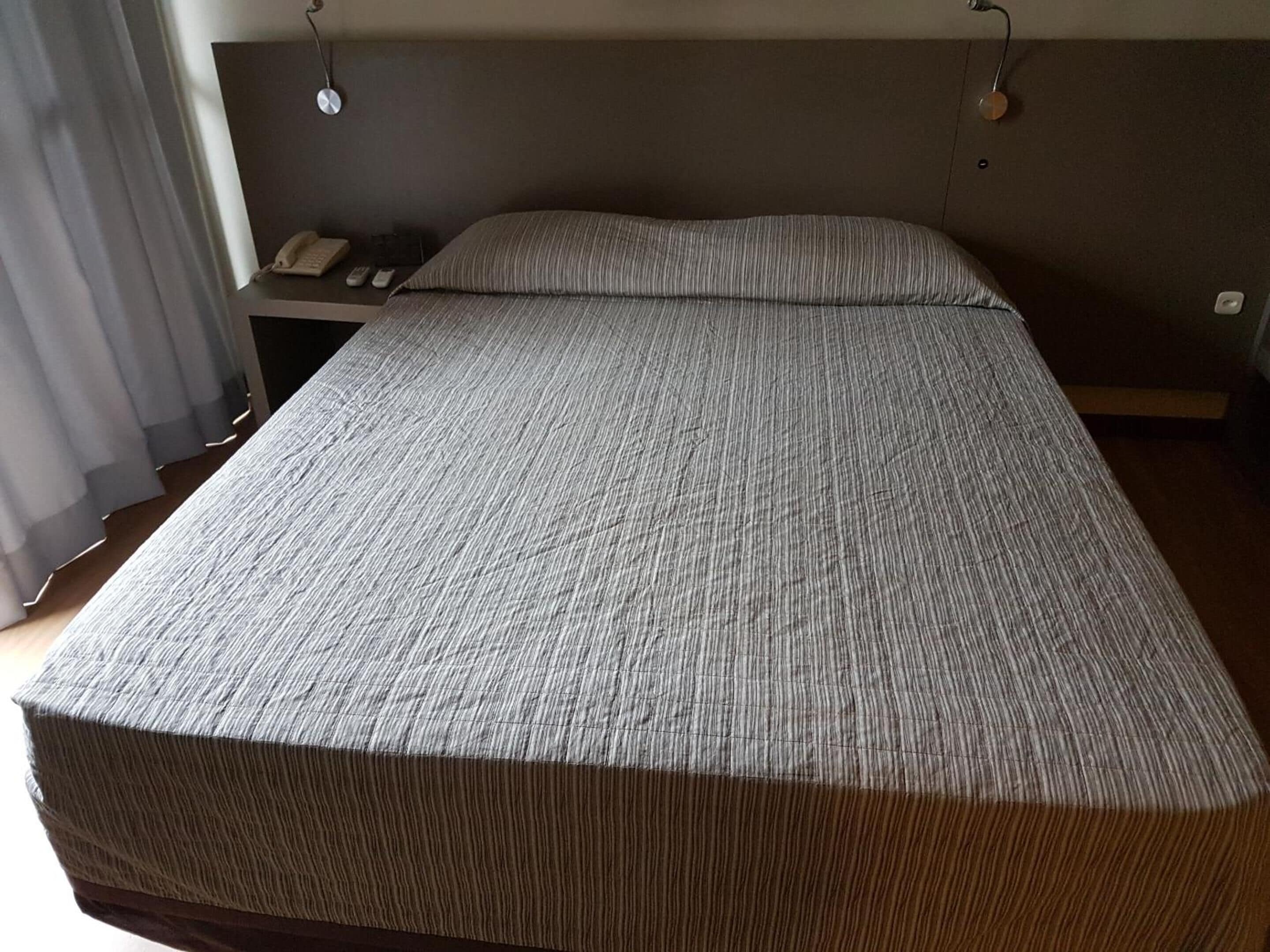 Una camera d'albergo con un letto grande, un comodino con telefono e telecomandi, e una tenda alla finestra.