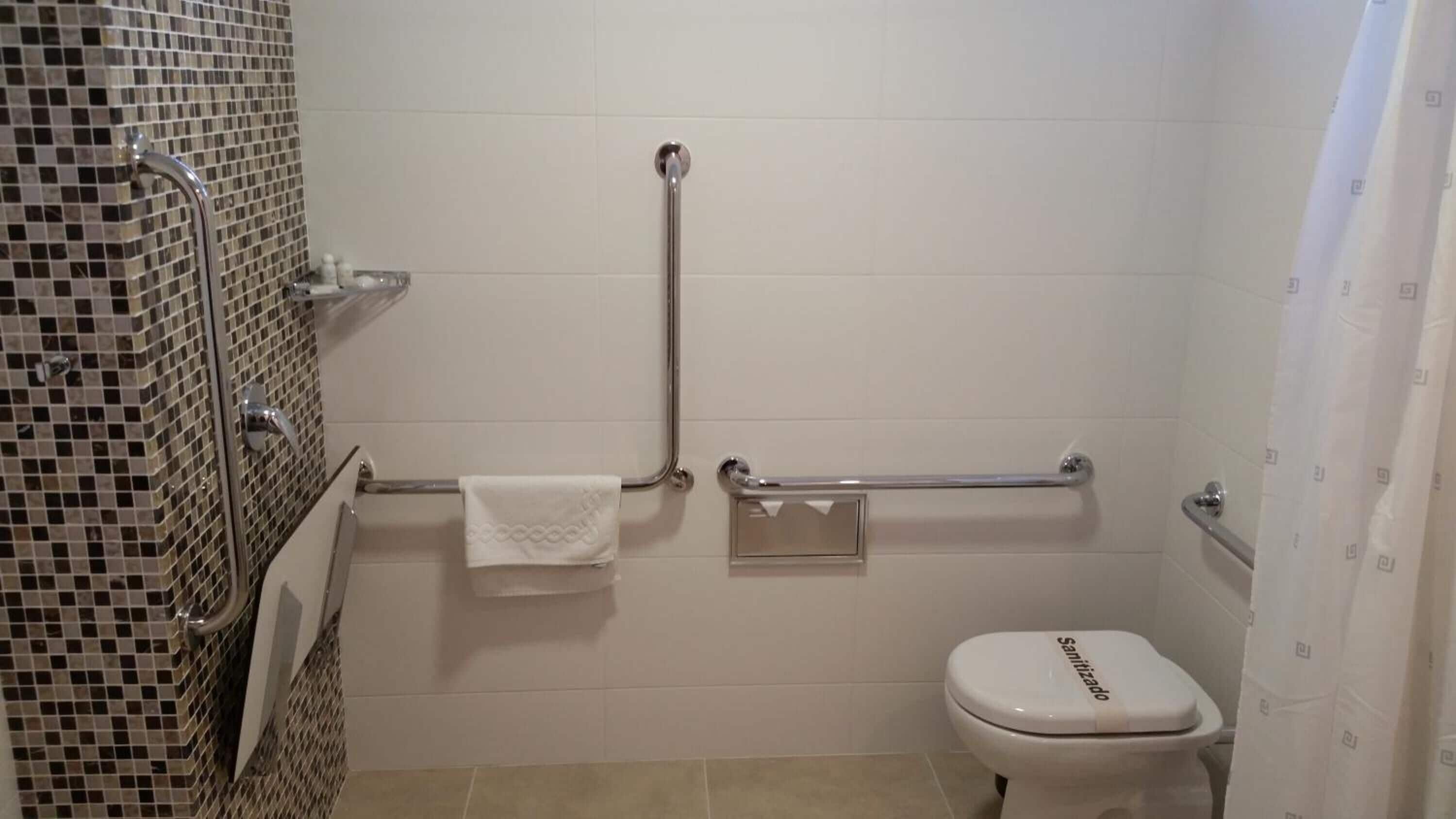 Un baño con inodoro blanco, barra de apoyo y pared con azulejos.