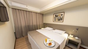 Una habitación de hotel con una cama, una mesita de noche con un teléfono y una taza, una bandeja con comida y bebida, y un cuadro en la pared.