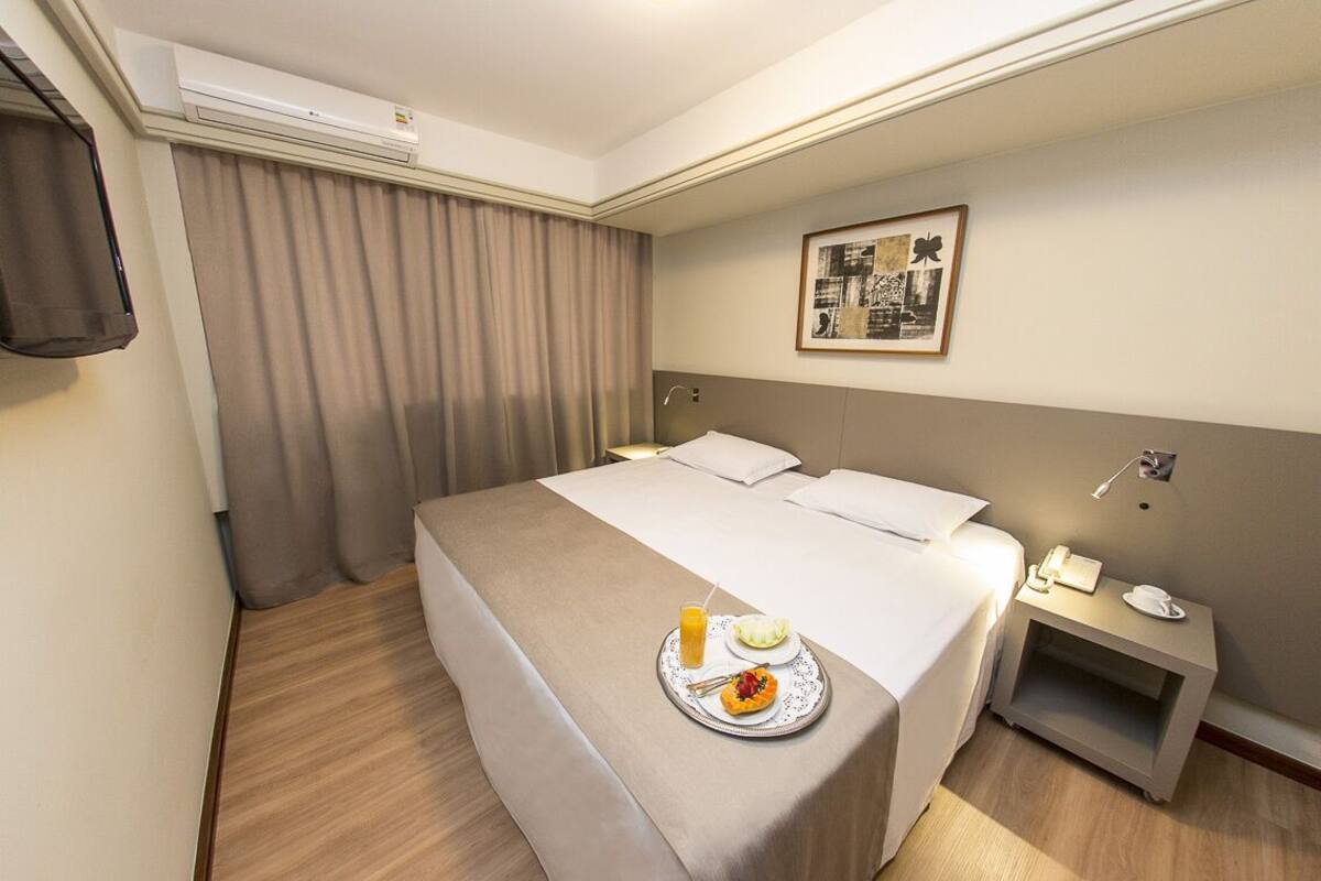 Hotelkamer met een bed, nachtkastje met telefoon en kopje, dienblad met eten en drinken, en schilderij aan de muur.
