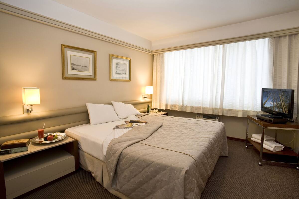 Hotelkamer met een groot bed, twee bedlampjes, een flatscreen televisie op een standaard, een raam met dunne gordijnen en twee ingelijste foto's aan de muur.