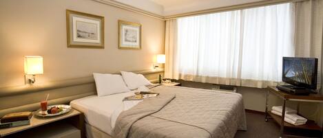 A hotel room with a large bed, two bedside lamps, a flat-screen TV on a stand, a window with sheer curtains, and two framed pictures on the wall.