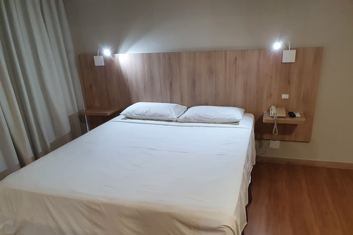 Een opgemaakt bed met witte lakens, een houten hoofdbord, bedlampjes en een telefoon op een klein plankje.