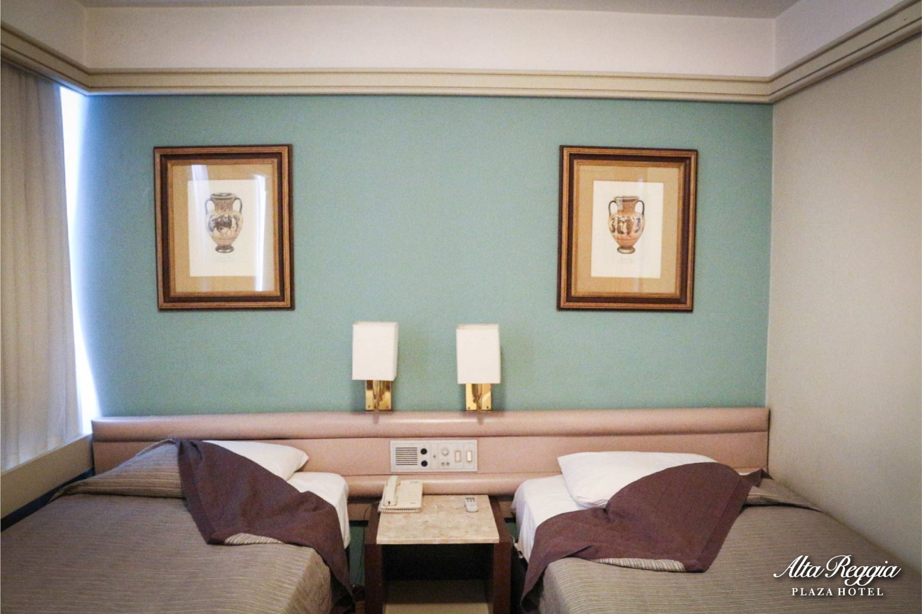 A hotel room with two beds, a small table with a telephone, and two wall-mounted lamps.