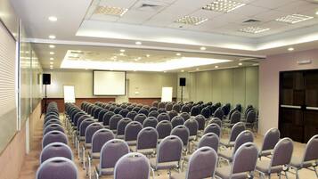 Uma sala de conferências com fileiras de cadeiras, uma tela de projetor e um palestrante.