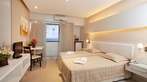 A hotel room with a bed, bedside table, desk, chair, and a view of the sea through the window. - Bahiamar Hotel (Salvador)