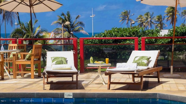 A poolside area with lounge chairs and umbrellas, a small table with a drink, and a view of palm trees and the ocean. - Bahiamar Hotel (Salvador)