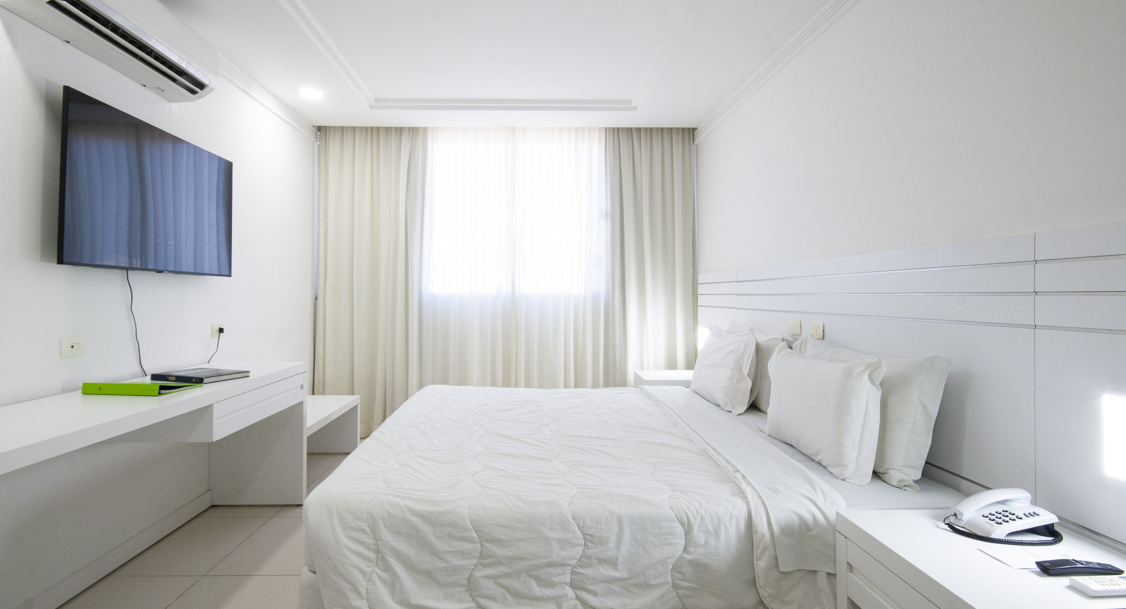 Une chambre d’hôtel équipée d’un lit, d’un bureau avec un ordinateur, d’un téléphone et d’un téléviseur fixé au mur.