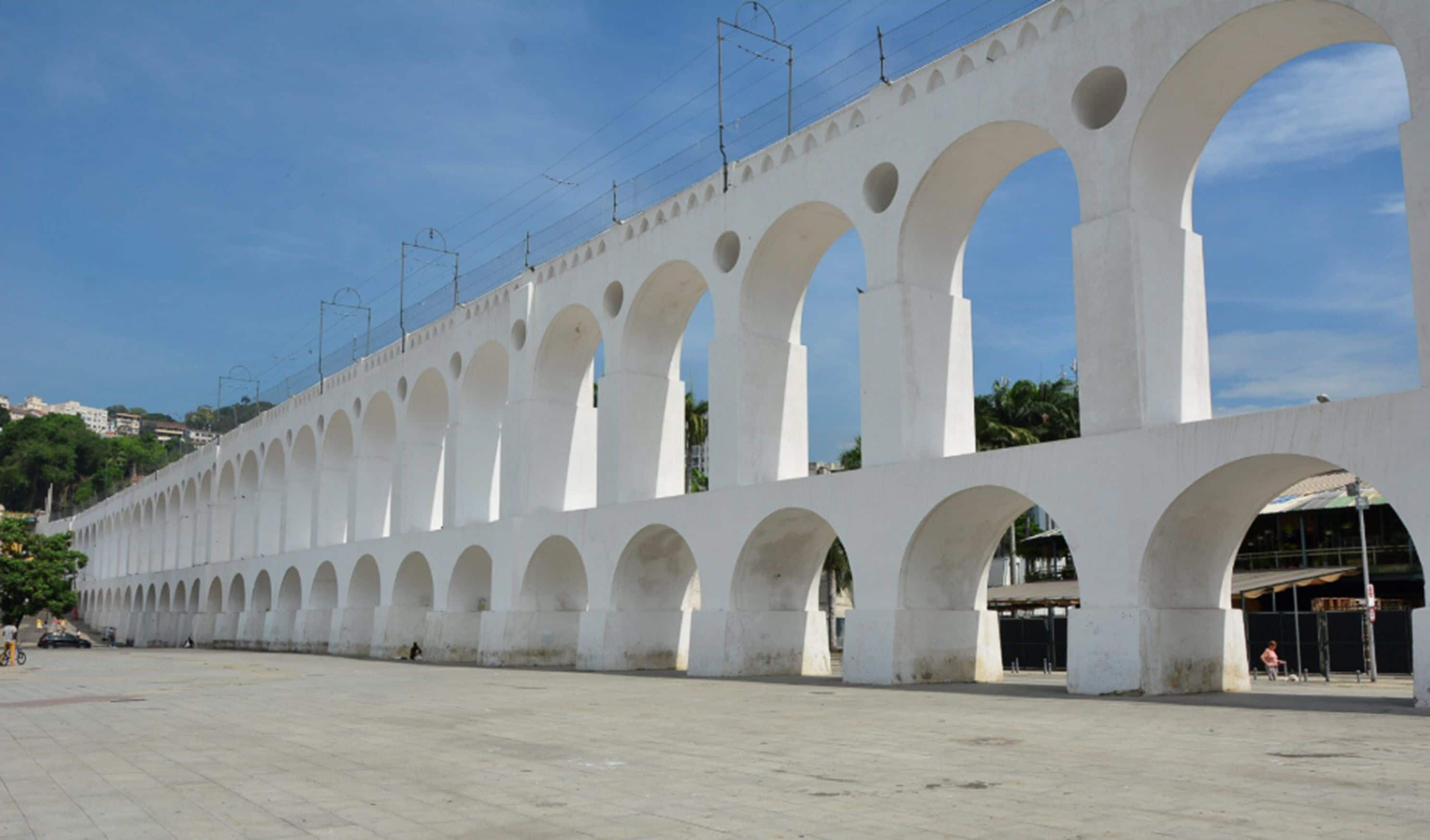A long white building with multiple arches and a flat roof, featuring a series of evenly spaced columns and a clear sky above.