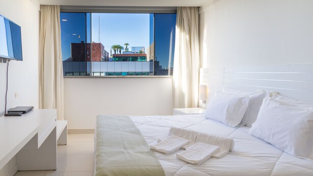A hotel room with a bed, a desk, a TV, and a window with a view of buildings and palm trees. - Hotel Bandeirantes (Rio de Janeiro)