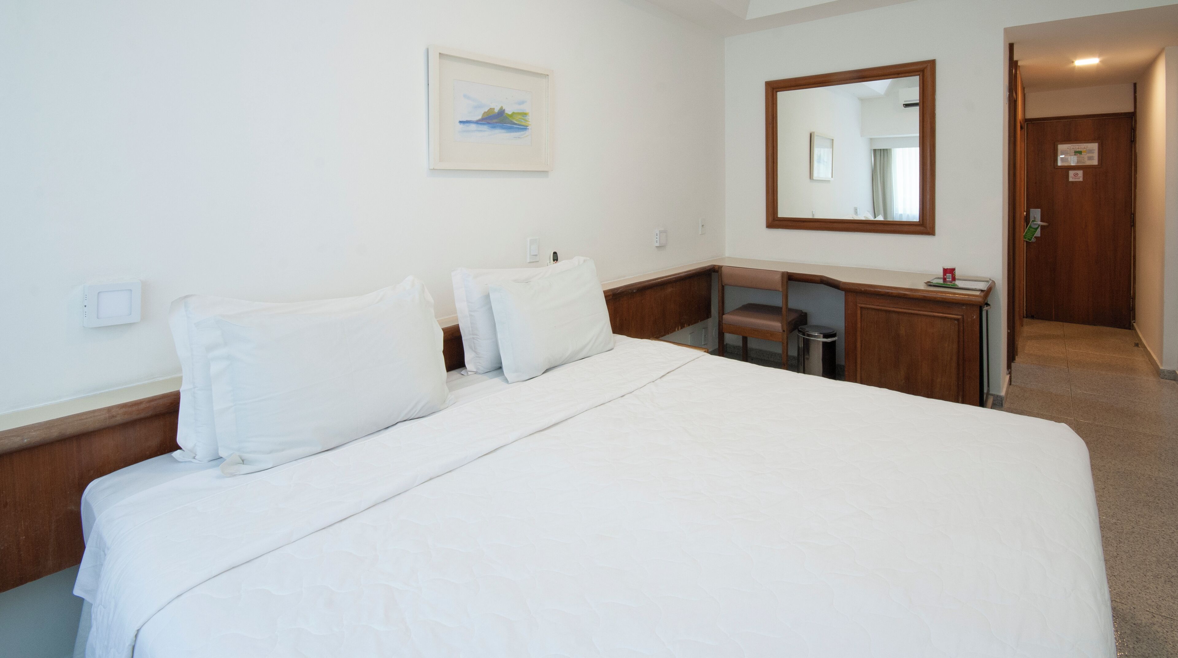Ein Hotelzimmer mit einem großen Bett, einem hölzernen Kopfteil, einem Schreibtisch, einem Stuhl, einem Spiegel und einem gerahmten Bild an der Wand.