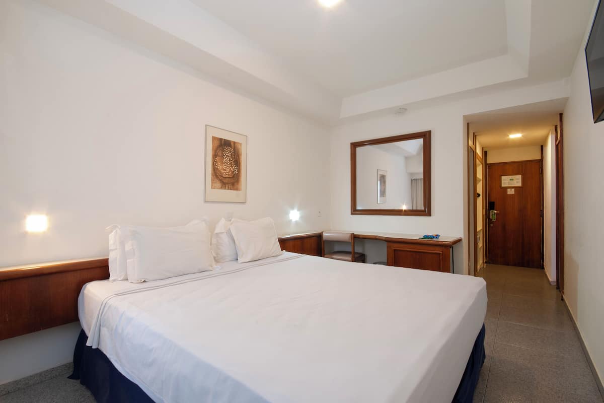 Una habitació d'hotel amb un llit gran, una tauleta de nit de fusta, un escriptori, un mirall i una porta que dona a una altra habitació.