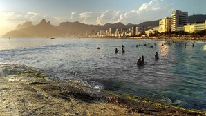 A beach with people swimming and sunbathing, a city skyline in the background, and mountains in the distance. - Hotel Bandeirantes (Rio de Janeiro)