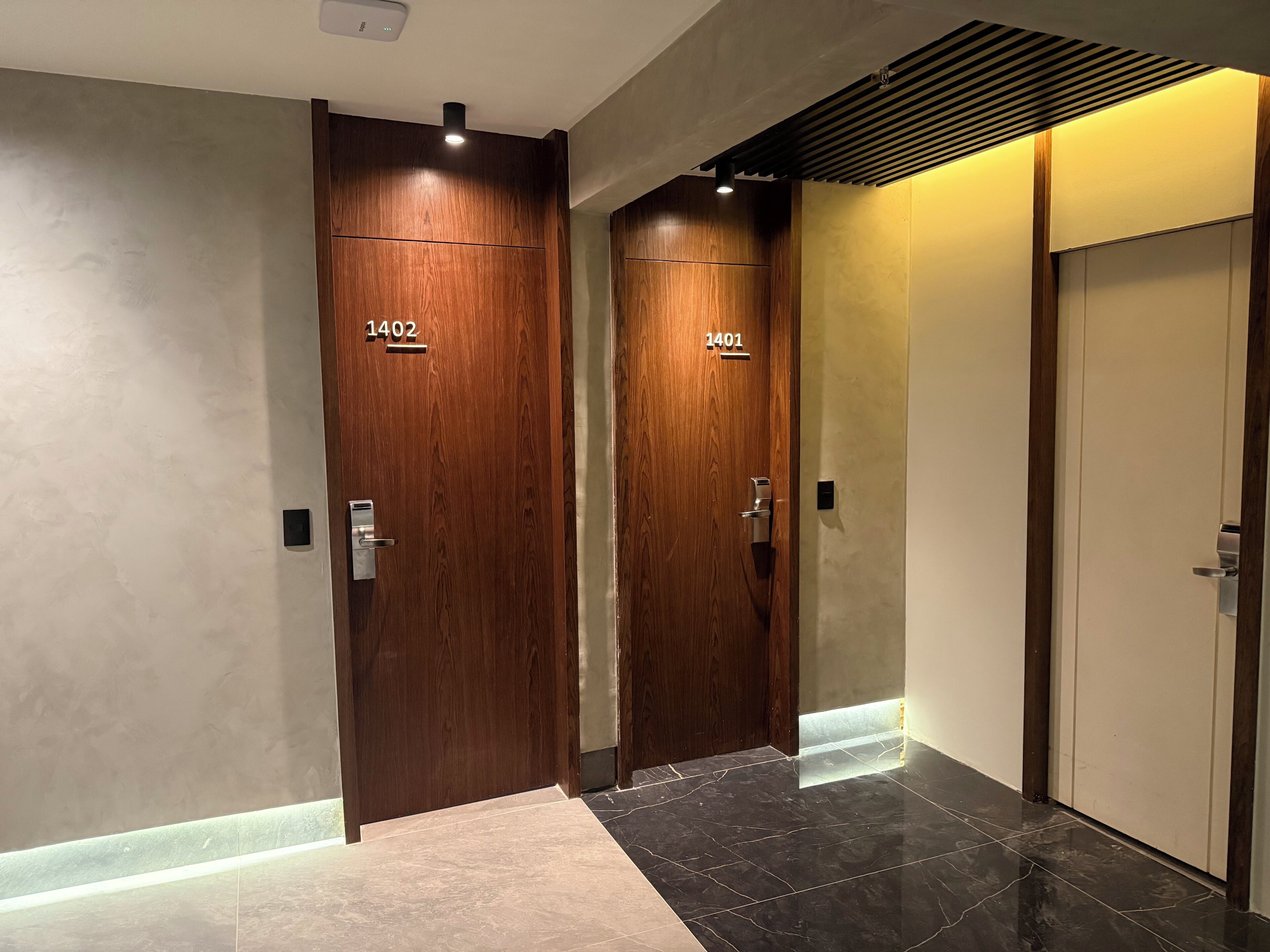 Deux portes en bois portant les numéros 1402 et 1401, un couloir moderne avec un sol carrelé et un plafond équipé d’un système de ventilation.