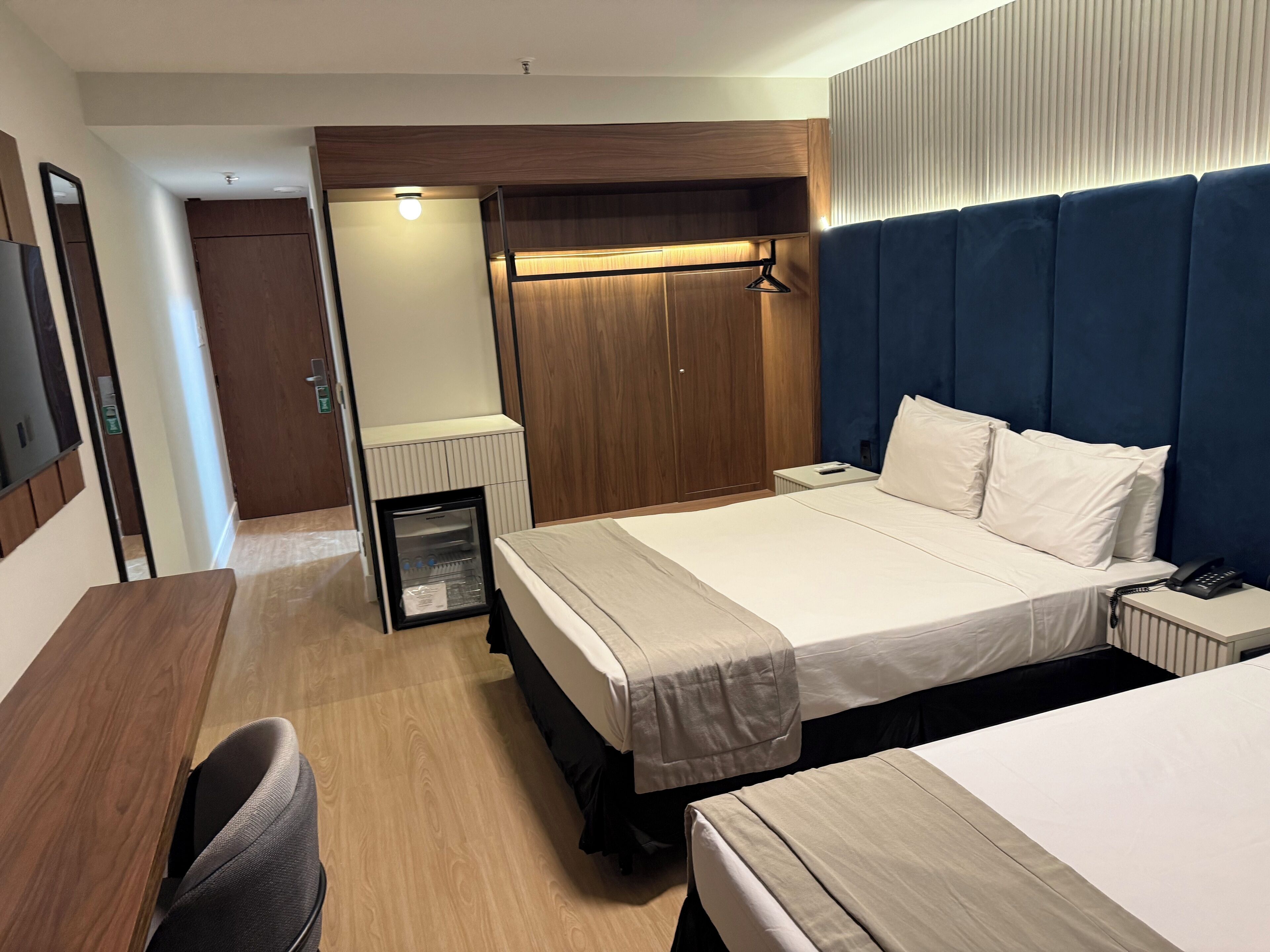 A hotel room with a bed, bedside table, chair, wardrobe, and a small refrigerator.