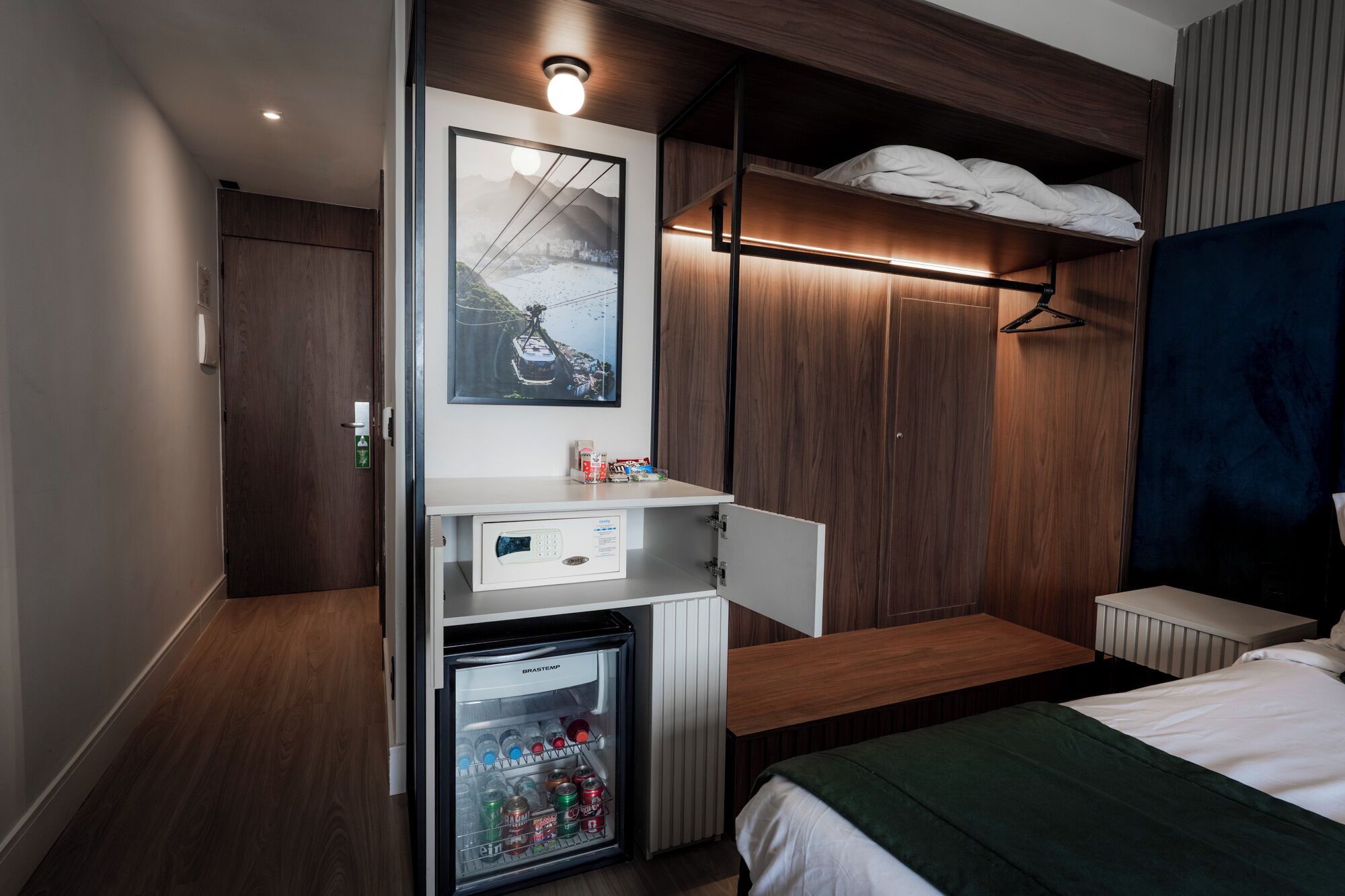 Habitación de hotel con cama, minibar, perchero y televisor en la pared.