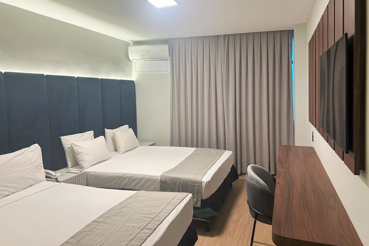 A hotel room with two beds, a desk, a chair, a TV, and an air conditioning unit.