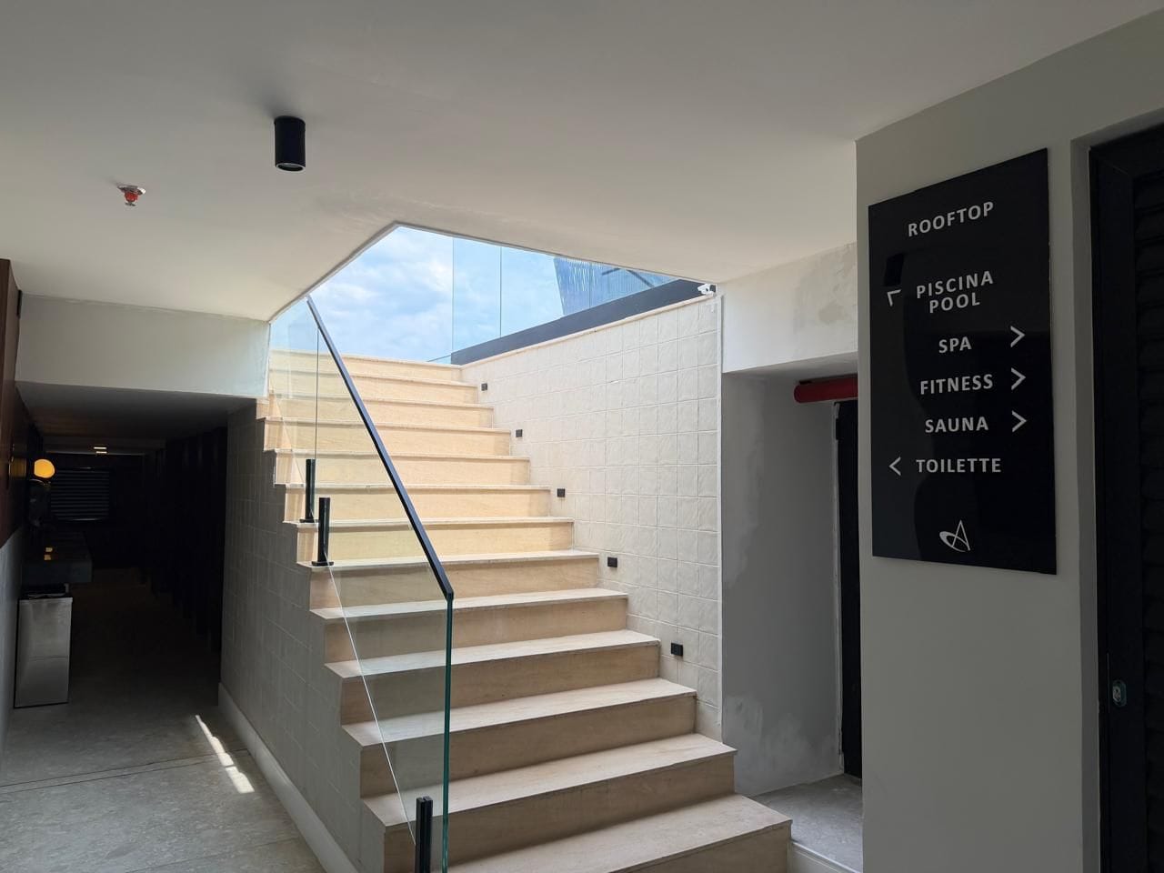 A modern staircase with a glass ceiling leading to a rooftop area. A sign indicates amenities including a pool, spa, fitness center, sauna, and toilet.