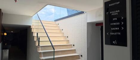 Eine moderne Treppe mit einer Glaskuppel, die zu einer Dachterrasse führt. Ein Schild weist auf Annehmlichkeiten wie Schwimmbad, Spa, Fitnessraum, Sauna und Toilette hin.
