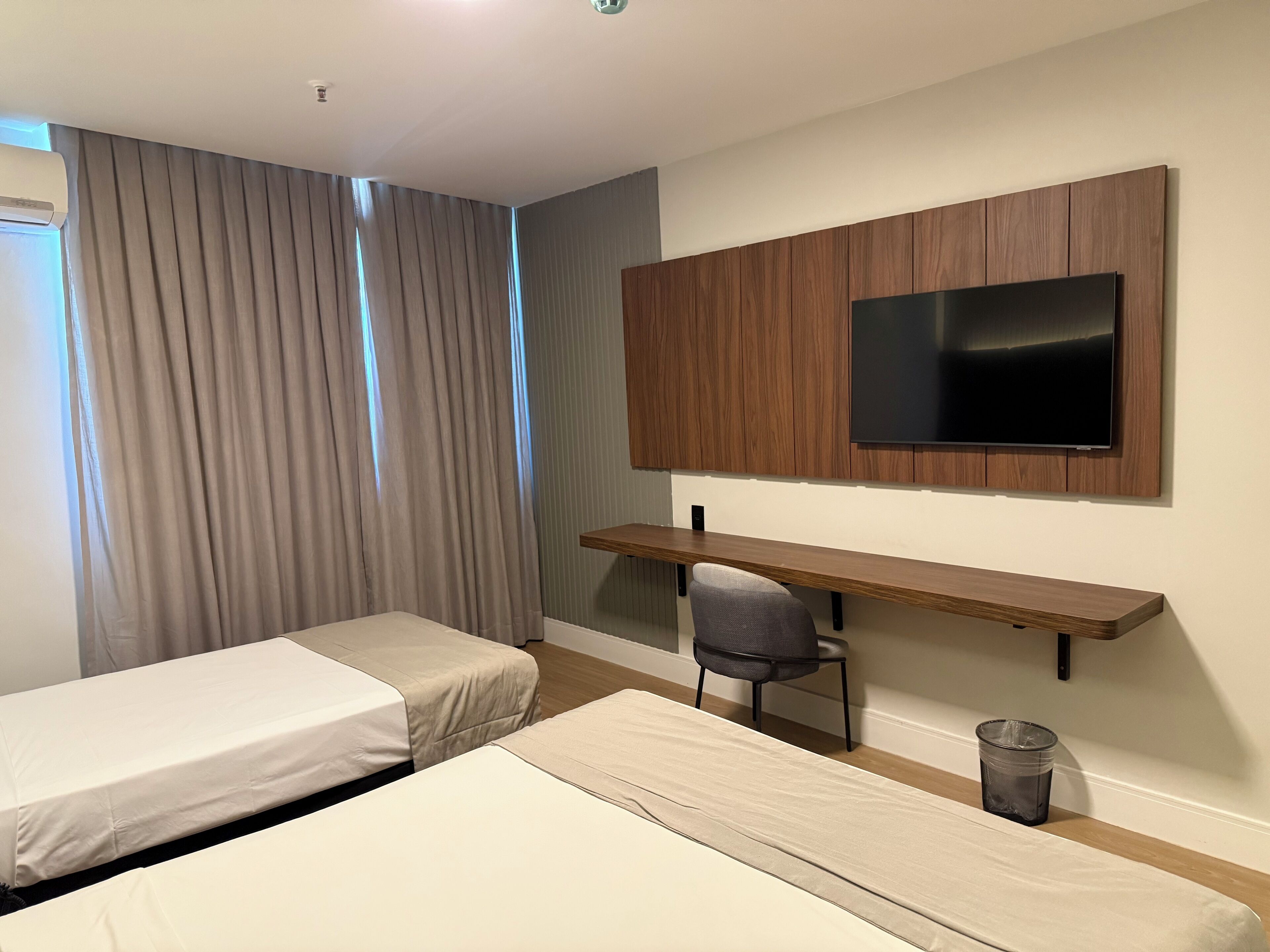 Een hotelkamer met twee eenpersoonsbedden, een bureau met stoel, een televisie bevestigd aan een houten paneel en een raam met gordijnen.