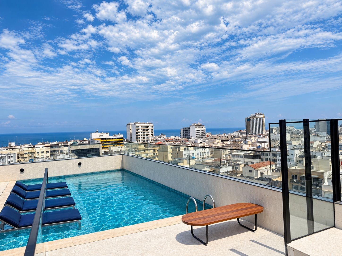 A rooftop pool with a bench and lounge chairs, overlooking a cityscape and the sea.
