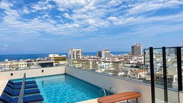 A rooftop pool with a bench and lounge chairs, overlooking a cityscape and the sea.