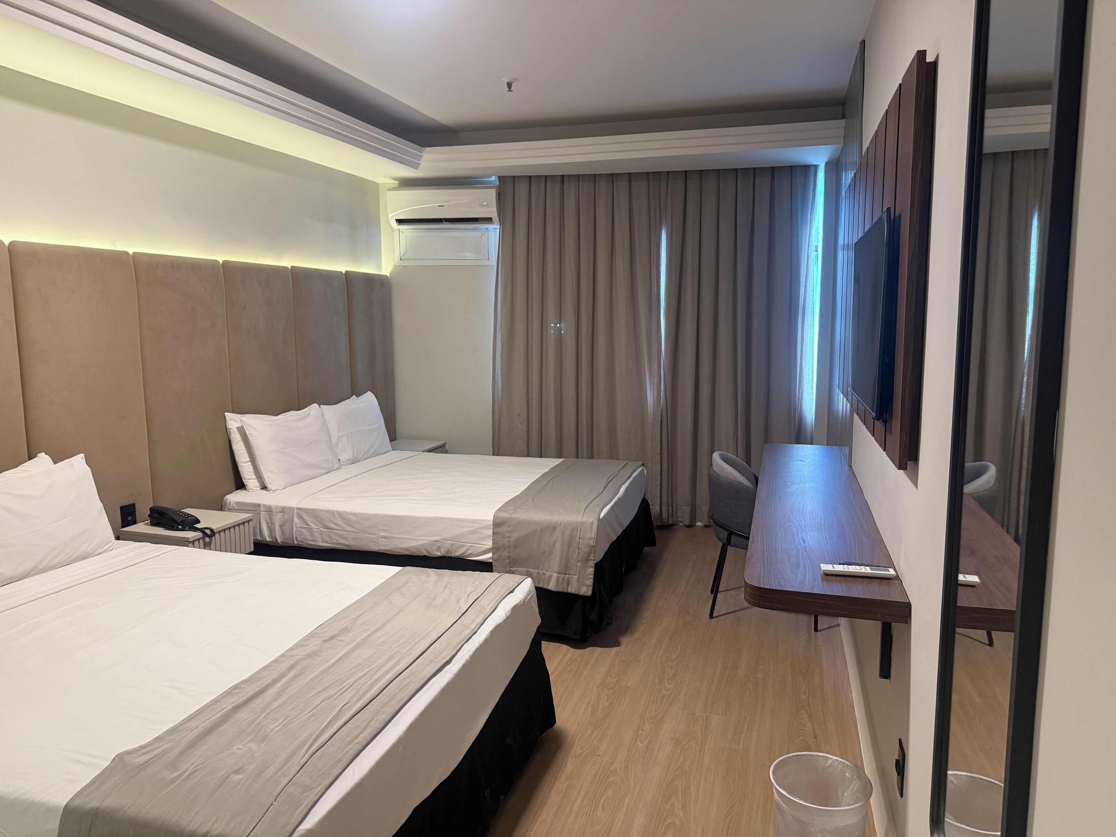 A hotel room with two beds, a desk, a chair, an air conditioning unit, and a large mirror.