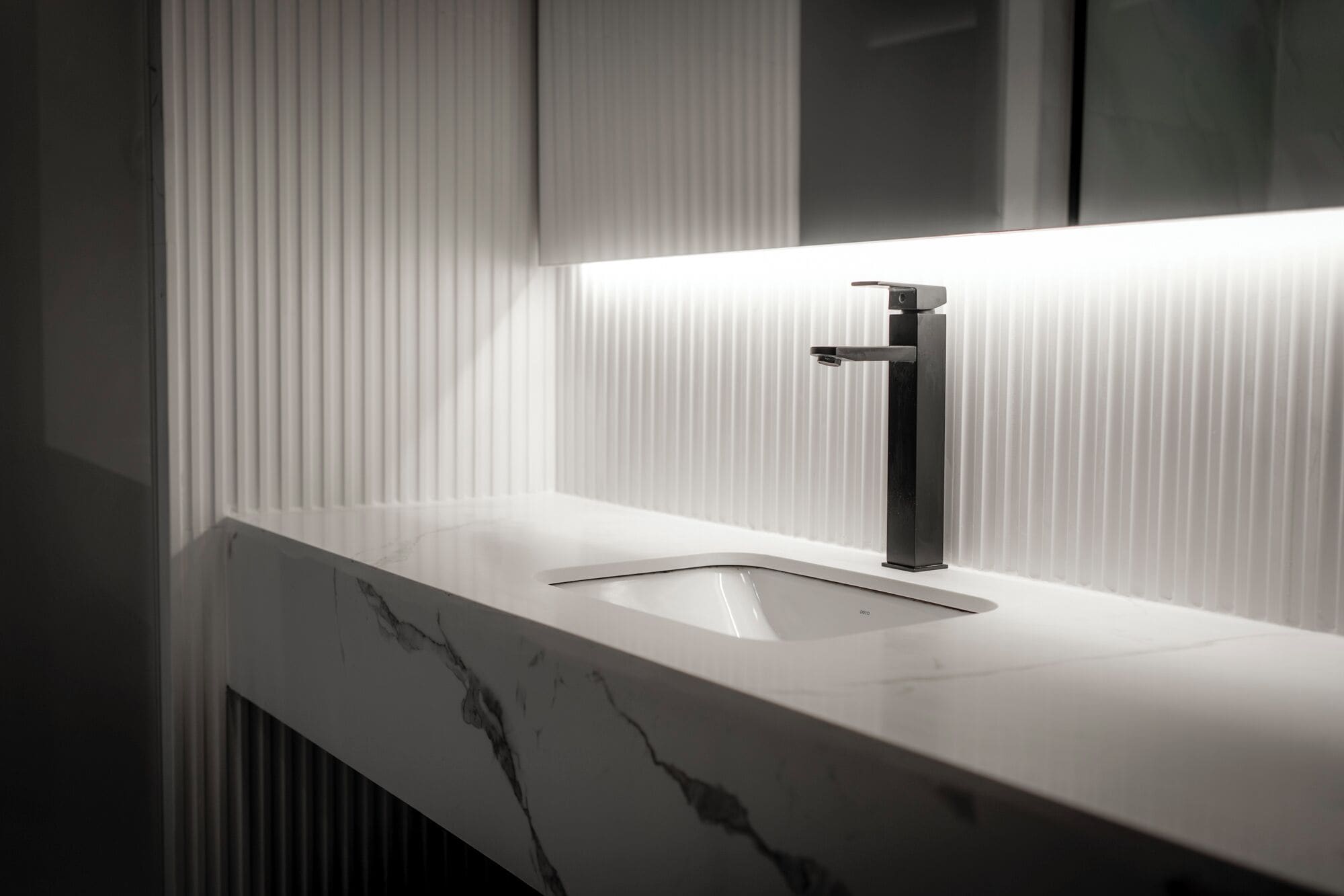 Un baño moderno con un elegante mostrador de mármol blanco, un lavabo individual y una pared con paneles verticales.