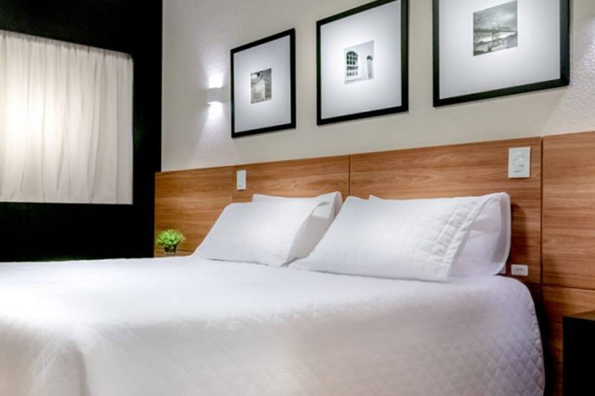 A neatly made bed in a hotel room with a wooden headboard, white bedding, and three framed pictures on the wall.
