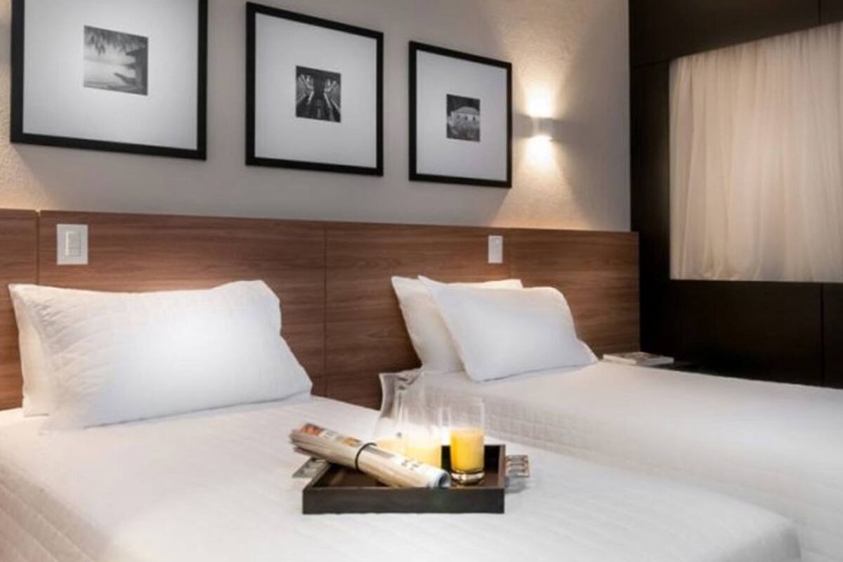 A hotel room with two beds, a headboard, framed pictures on the wall, and a tray with a newspaper, glasses, and a bottle.
