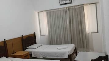Quarto de hotel com duas camas de solteiro, uma mesa de cabeceira de madeira, uma pequena mesa, uma cadeira e um aparelho de ar condicionado na parede.
