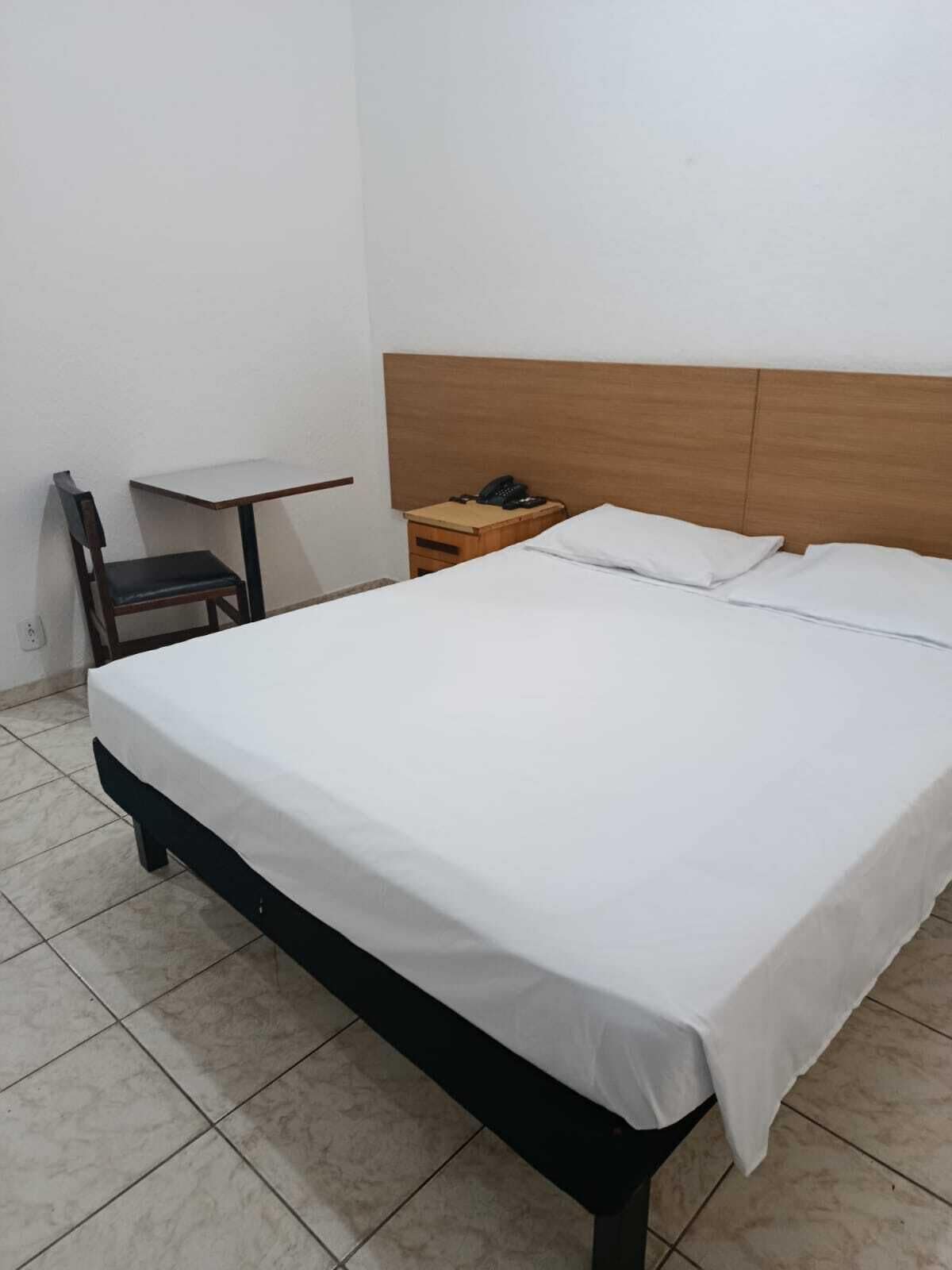 A single bed with a wooden headboard, a small wooden table, and a chair in a room with tiled flooring.