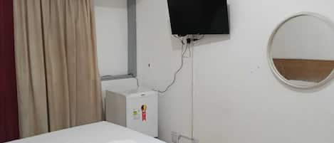Habitación de hotel con cama, televisor montado en la pared, unidad de aire acondicionado pequeña, espejo y cortina.