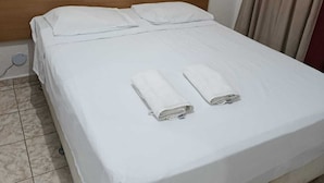 Una cama bien hecha con sábanas blancas y dos toallas enrolladas, un cabecero de madera y una cortina abierta a un lado.