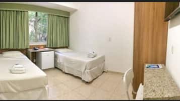 Quarto pequeno e limpo com duas camas, uma escrivaninha e uma janela com cortinas.